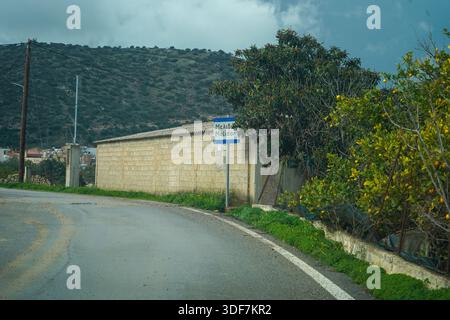 Un cartello stradale che segna l'ingresso al paese di Melidoni, Creta. Circondato da una lussureggiante vegetazione verde, alberi di agrumi e da un tradizionale muro di pietra Foto Stock