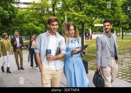 Gruppo di diversi professionisti e imprenditori che camminano lungo un sentiero lastricato in un parco urbano verde, discutendo di lavoro in collaborazione e di utilizzo Foto Stock