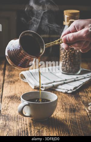 Versare il caffè turco in una tazza bianca. Vasetto di chicchi di caffè tostati su sfondo Foto Stock