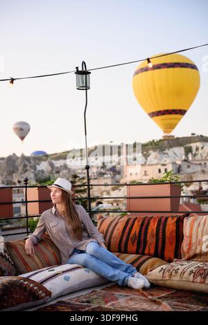 Una donna rilassata si gode l'alba della Cappadocia mentre si siede sui cuscini sulla terrazza dell'hotel, guardando le mongolfiere e la valle rocciosa la mattina presto. Il viaggiatore sorride ammirando la pittoresca valle Foto Stock