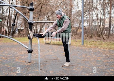 Un uomo con tuta e cappello si allunga su un campo sportivo all'aperto. Esercizio fisico a basse temperature. Foto Stock
