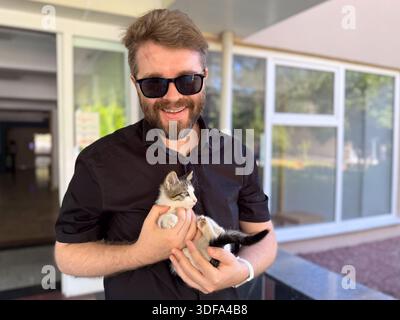 Uomo con occhiali da sole sorridente e in mano un piccolo gattino all'aperto. Compagnia affetto e legame emotivo tra uomo e animale. Foto Stock
