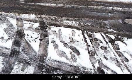 Footprints and tire tracks in melting snow on a wet urban street during winter Foto Stock