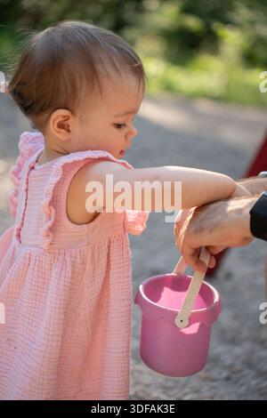 il bambino in abito rosa si impegna con un adulto che tiene in mano il secchio rosa nelle giornate di sole, catturando un sano momento di legame all'aperto con il morbido dorso verde e grigio Foto Stock
