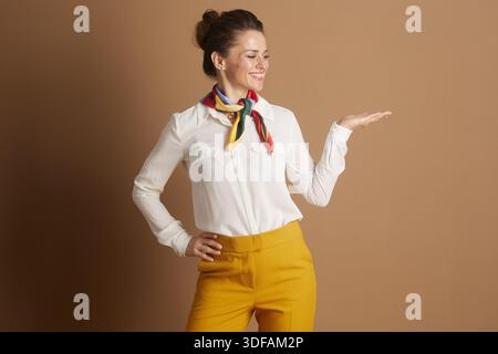 Una donna sorridente di mezza età, vestita professionalmente con una camicia bianca e pantaloni gialli senape con una sciarpa colorata, presenta o offre con sicurezza Foto Stock