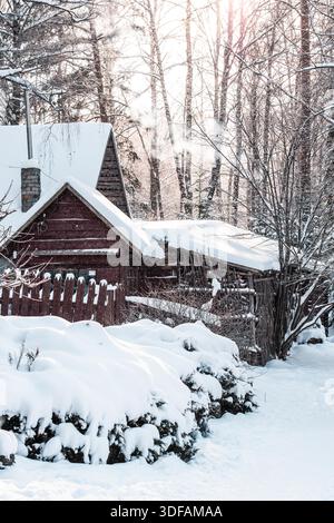 Case o capanne in legno coperte di neve con una recinzione rustica incastonata tra gli alberi in una tranquilla foresta invernale Foto Stock