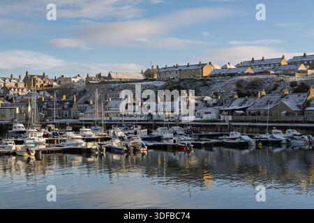 Il porto di Findochty è pieno di barche coperte di neve. Il sole splende sull'acqua, creando una scena luminosa. Le case lungo la riva si aggiungono a t Foto Stock