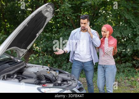 Triste despaired donna araba in hijab e l'uomo chiamata al supporto auto, stare vicino a macchina rotta con cofano aperto Foto Stock