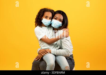 Ritratto di madre e figlia afroamericana che indossano maschere mediche protettive sul viso su sfondo giallo Foto Stock