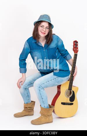 Felice sorridente giovane musicista donna seduta ordinatamente su una chitarra verticale e guarda la fotocamera. Donna di Zoomers, con occhiali da vista, vestita Foto Stock