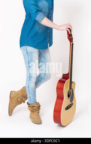 La musicista femminile irriconoscibile in denim casual si staglia con la chitarra classica tenuta verticalmente di fronte a lei. Giovane chitarrista vestita Foto Stock