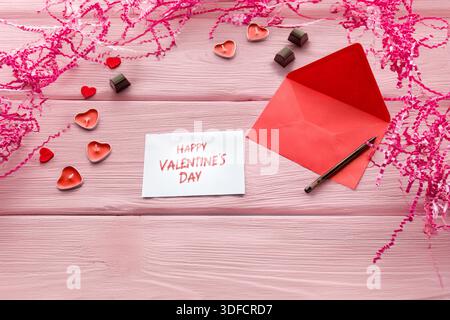 Strato piatto di buste rosse con cartoncino vuoto, penna, candele a cuore e cioccolatini su sfondo rosa. Foto Stock
