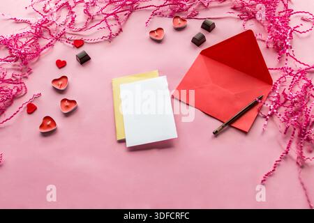 Strato piatto di buste rosse con cartoncino vuoto, penna, candele a cuore e cioccolatini su sfondo rosa. Foto Stock