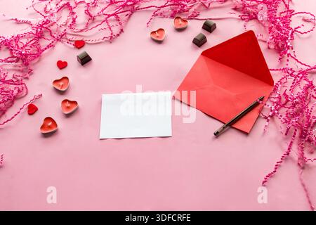 Strato piatto di buste rosse con cartoncino vuoto, penna, candele a cuore e cioccolatini su sfondo rosa. Foto Stock