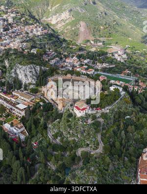 Vista aerea dell'antico Teatro Antico, immerso nel verde e nei sentieri tortuosi, contrasta con i tetti in terracotta della città, Taormina, S. Foto Stock