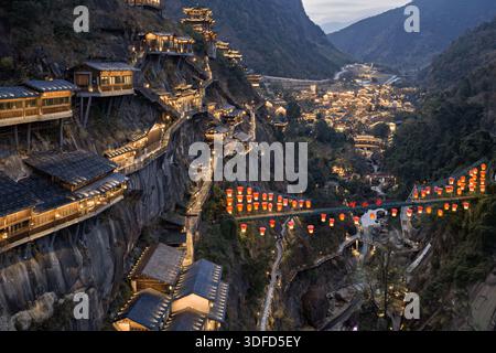 Vista aerea di case in legno arroccate su ripide scogliere, ponti illuminati adornati da lanterne rosse che emettono un caldo bagliore sulla Wangxian Valley, Wangxia Foto Stock