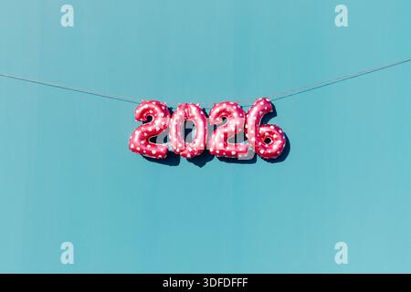 i palloncini a forma di cuore rosa che formano il numero 2026, come il nuovo anno, sono appesi da una sottile corda rossa contro una luminosa parete color ottanio, in un set da esterno pulito Foto Stock