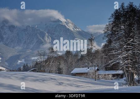 Berchtesgaden, Deutschland 11.01.2026: IM Bild: Verschneite Winterlandschaft mit Blick auf das alpine Bergmassiv. Von links sind der Hohe Göll sowie das Hohe Brett zu sehen, deren Gipfel teilweise von tief hängenden Wolken umgeben sind. Im Vordergrund liegen eine schneebedeckte Wiese, einzelne Bäume und Gebäude unter klarem Winterhimmel. Bayern *** Berchtesgaden, Germania 11 01 2026 nella foto paesaggio invernale innevato con vista sul massiccio alpino da sinistra si possono vedere gli alti Göll e gli alti Brett, le cui vette sono in parte circondate da basse nuvole in primo piano Foto Stock
