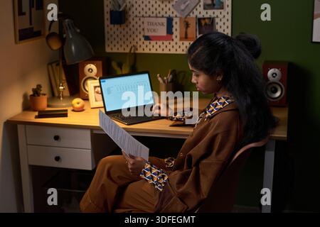 Giovane donna dell'Asia meridionale che esamina le carte stampate mentre è seduta alla scrivania Foto Stock