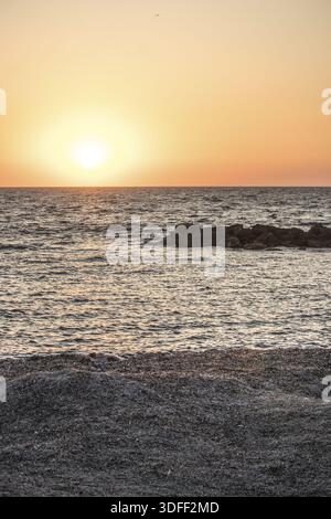 Vista del tramonto sul mare tranquillo e scintillante, che getta un caldo bagliore dorato sulle rocce scure e sulla spiaggia sabbiosa di Paphos, Paphos e Cipro. Foto Stock
