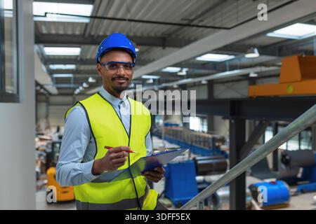 Un manager maschile supervisiona i lavoratori di uno stabilimento di produzione promuovendo il lavoro di squadra e la comunicazione nelle operazioni industriali. Foto Stock