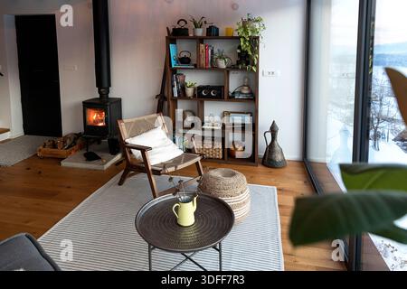 Intimo soggiorno interno in stile scandinavo con caminetto e finestra panoramica con mobili minimalisti, piante e vista panoramica sulla campagna Foto Stock