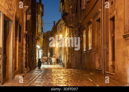 Vista della strada acciottolata inondata di luce calda, fiancheggiata da edifici antichi, mentre figure sfocate vagano in lontananza, Rovigno, Istria County, CR Foto Stock