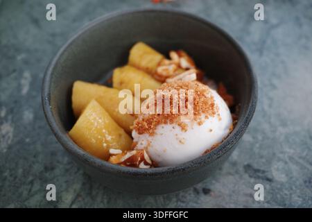 Una deliziosa ciotola da dessert con banane caramellate gialle dolci servite con una paletta di cremoso gelato bianco al cocco, guarnito con cru tostato Foto Stock