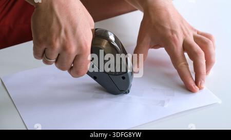 Utilizzando un dispenser di scotch. Incollare del nastro su un pezzo di carta. Formazione sull'uso della cancelleria. Pezzi di nastro abrasivo. Primo piano con le mani di un uomo che lavora Foto Stock