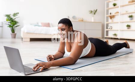 Donna nera sovrappeso sdraiata su un tappetino sportivo, utilizzando un computer portatile, comunicando con un allenatore personale o guardando un video di fitness Foto Stock