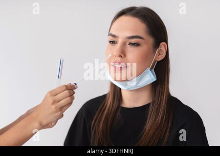 Medico che fa covid-19 test PCR nasale per paziente giovane femmina su sfondo studio chiaro Foto Stock
