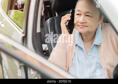 Donna asiatica anziana che usa il telefono e apre la porta della macchina. Donna anziana che esce dall'auto mentre parla con lo smartphone. Foto Stock