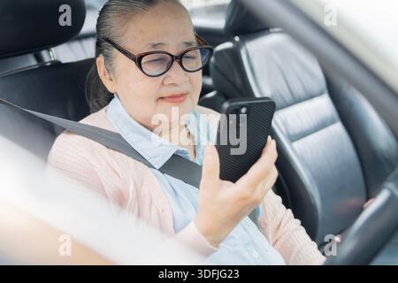 Donna anziana sorridente che tiene in mano lo smartphone mentre è seduta in auto, donna matura che guida l'auto e utilizza il telefono. Concetto di stile di vita pensionistico. Foto Stock