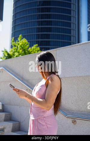Giovane donna d'affari che indossa gli occhiali da sole usando lo smartphone all'aperto vicino a un moderno edificio di uffici Foto Stock