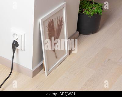 Una cornice bianca racchiude un ramo di pino secco, appoggiato su un pavimento di legno chiaro accanto a uno sbocco e a una pianta, suggerendo la bellezza della natura in un ambiente domestico. Foto Stock