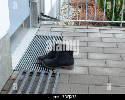 Un paio di stivali Chelsea neri poggia su una griglia di metallo vicino a un patio piastrellato, suggerendo l'uso all'aperto, pratiche calzature quotidiane e la preparazione per il generale Foto Stock
