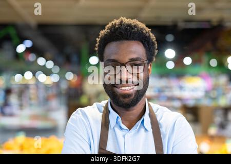 Giovane uomo afroamericano adulto con un sorriso caldo e con un grembiule, che lavora in un negozio di alimentari, rappresentando il servizio clienti e l'ospitalità Foto Stock
