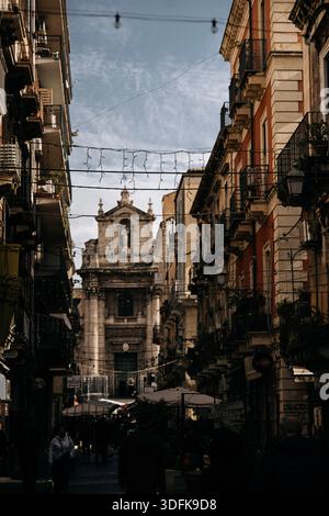 Strada stretta a Catania con facciata barocca della chiesa: Scena urbana italiana siciliana incorniciata da alti edifici storici e cavi sopraelevati. Alta qualità Foto Stock
