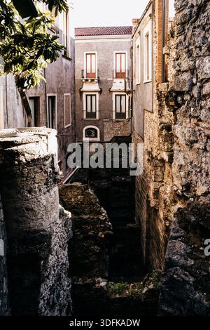 Teatro Romano e Odeon di Catania, Sicilia: Antiche rovine del teatro romano con posti a sedere in pietra, edifici storici e torri della chiesa sotto la luce del sole. Foto Stock