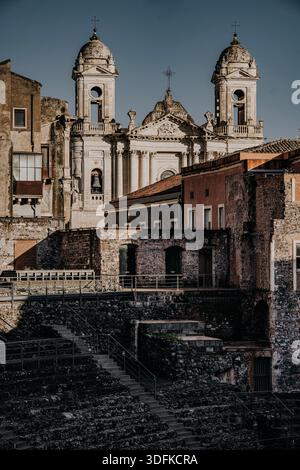 Teatro Romano e Odeon di Catania, Sicilia: Antiche rovine del teatro romano con posti a sedere in pietra, edifici storici e torri della chiesa sotto la luce del sole. Foto Stock