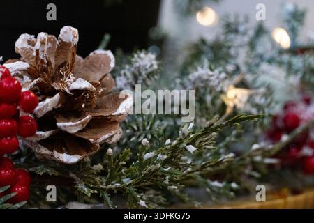 Decorazioni natalizie natalizie con bacche rosse, cono di pino e rami sempreverdi ricoperti di neve artificiale. Accogliente atmosfera di vacanza con luci calde, Foto Stock