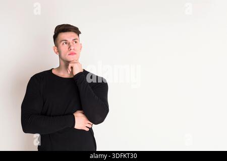 Giovane uomo che ha riflettuto attentamente su una parete bianca semplice indossando una camicia a manica lunga scura Foto Stock