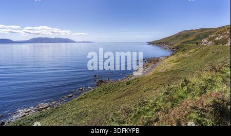 Blue sky with white puffy clouds over rocky costal line and mountains in a distance Foto Stock