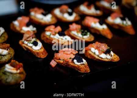 I pezzi di pane tostato ricoperti da formaggio spalmabile, cubetti di salmone e caviale nero sono esposti su un vassoio scuro Foto Stock