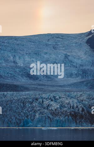 Vista di un colossale ghiacciaio, la sua distesa blu ghiacciata che incontra le acque tranquille sotto un bagliore morbido ed etereo, Fjallsarlon, ghiacciaio Vatnajokull, Islanda. Foto Stock
