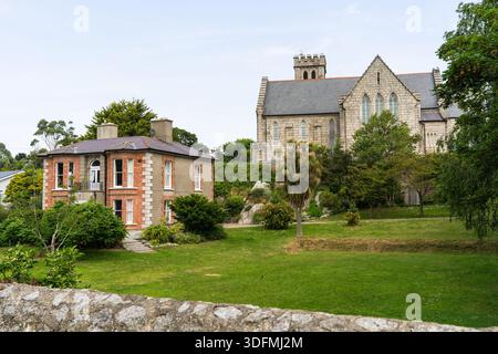 Vista della vecchia casa in mattoni con finiture bianche e una grande chiesa in pietra con una torre su una collina circondata da erba verde e alberi, Dalkey, Dublino, Irlanda. Foto Stock