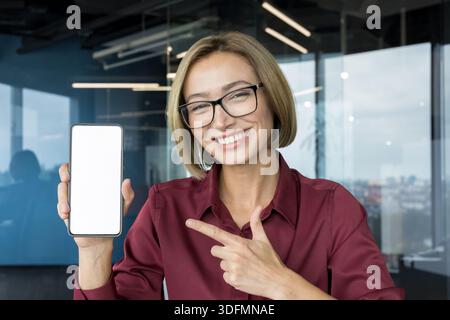Donna d'affari sorridente con occhiali e camicia Bordeaux che punta a uno smartphone con schermo bianco vuoto, che presenta un mockup di app mobile in un ambiente d'ufficio moderno, sicura di sé e professionale Foto Stock