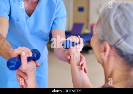 Una donna anziana sta facendo esercizi al braccio con manubri blu durante una sessione di fisioterapia con un terapista maschile in un centro di riabilitazione al chiuso. Questa immagine c Foto Stock