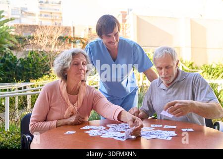 Una coppia senior gioca a carte con un fisioterapista all'aperto durante una sessione di terapia riabilitativa presso un centro di riabilitazione che si concentra sull'allenamento cerebrale e sulla c Foto Stock