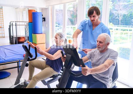 I pazienti anziani utilizzano cyclette con un fisioterapista che li supervisiona in un centro di riabilitazione per il recupero e la riabilitazione al chiuso. Questa immagine Foto Stock